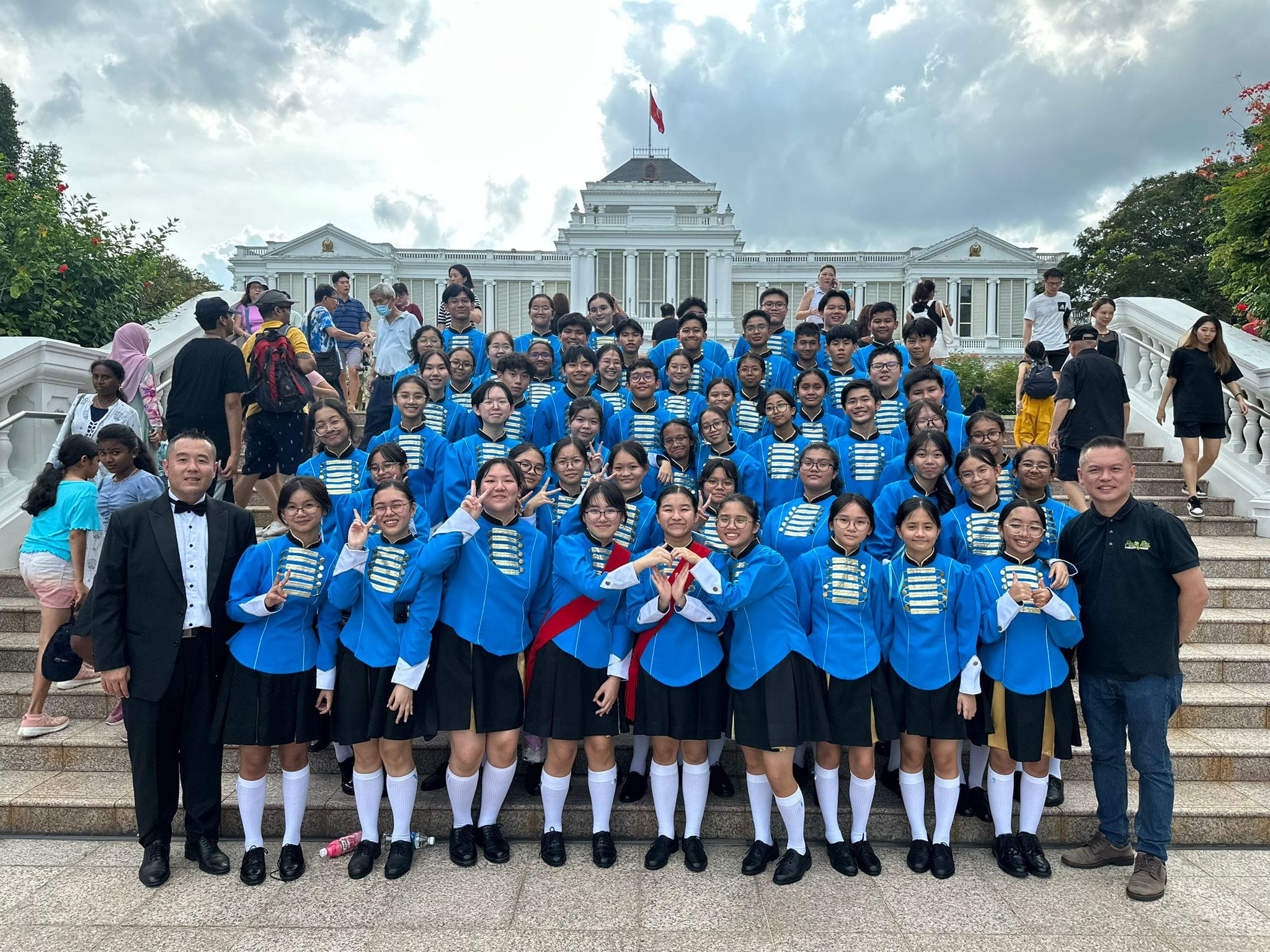 A large group of youth in blue marching band uniforms poses on steps in front of a white colonial building with a red flag.